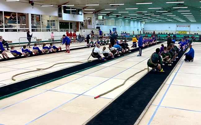 Two tug of war teams pulling on ropes laid down matting across the boarded floor, with spectators along the sides