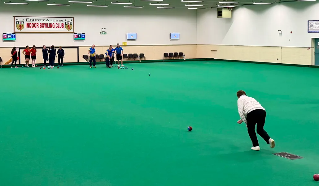 A bowler in delivery on the green, with players waiting at the far end beneath the club banner